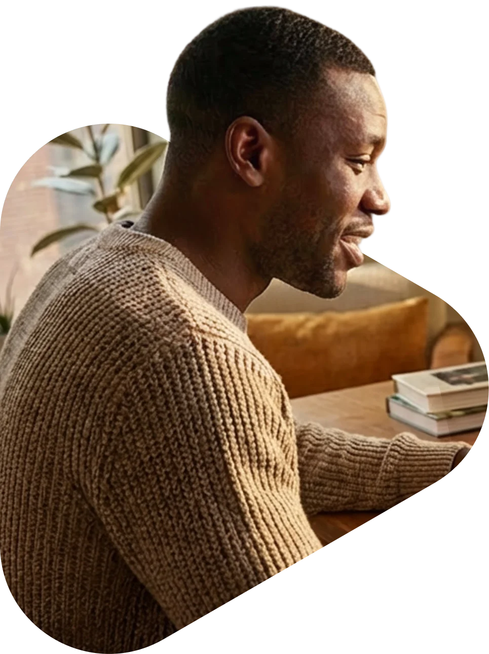 A man in a brown knit sweater sitting at a wooden table, talking, with books and a cozy living room in the background.